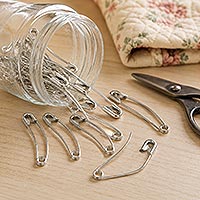 A glass jar of curved safety pins spilled over beside folded fabric and scissors.