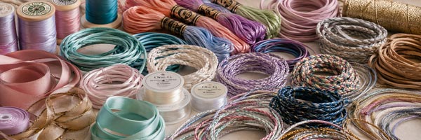 Colorful assortment of craft cords, ribbons, threads, and jewelry stringing materials including satin ribbon, braided cords, metallic cords, beading thread, and decorative fibers.